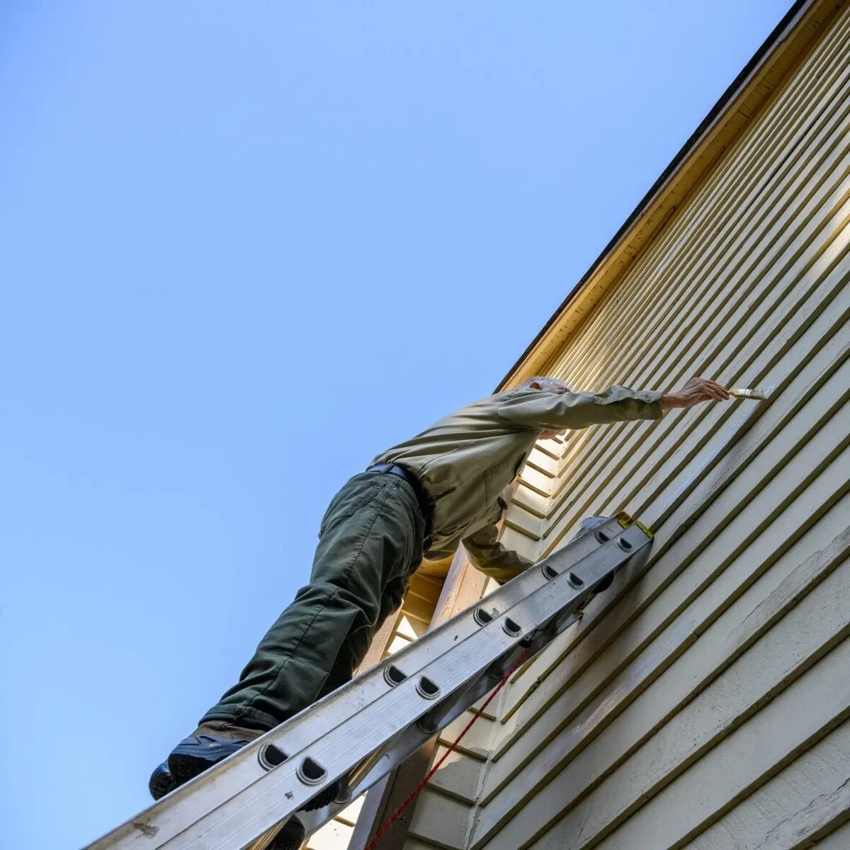 A person on a ladder painting the beige siding of a house with a brush, against a clear blue sky.