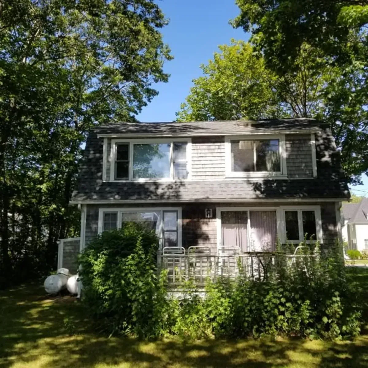 A gray, two-story house stands surrounded by lush green trees and a well-maintained lawn.