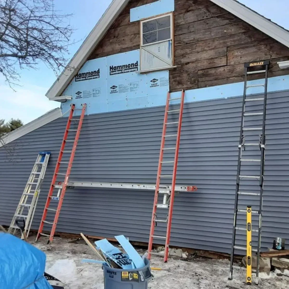 Construction workers install blue siding on a house with two ladders leaning against it.