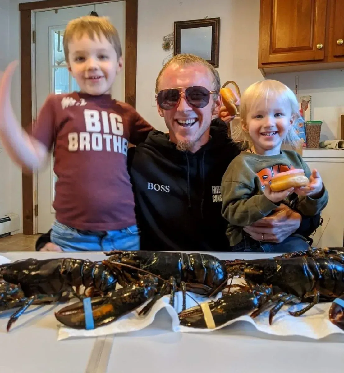 A smiling man in sunglasses with two happy children, one wearing a 'BIG BROTHER' shirt, and several live lobsters.