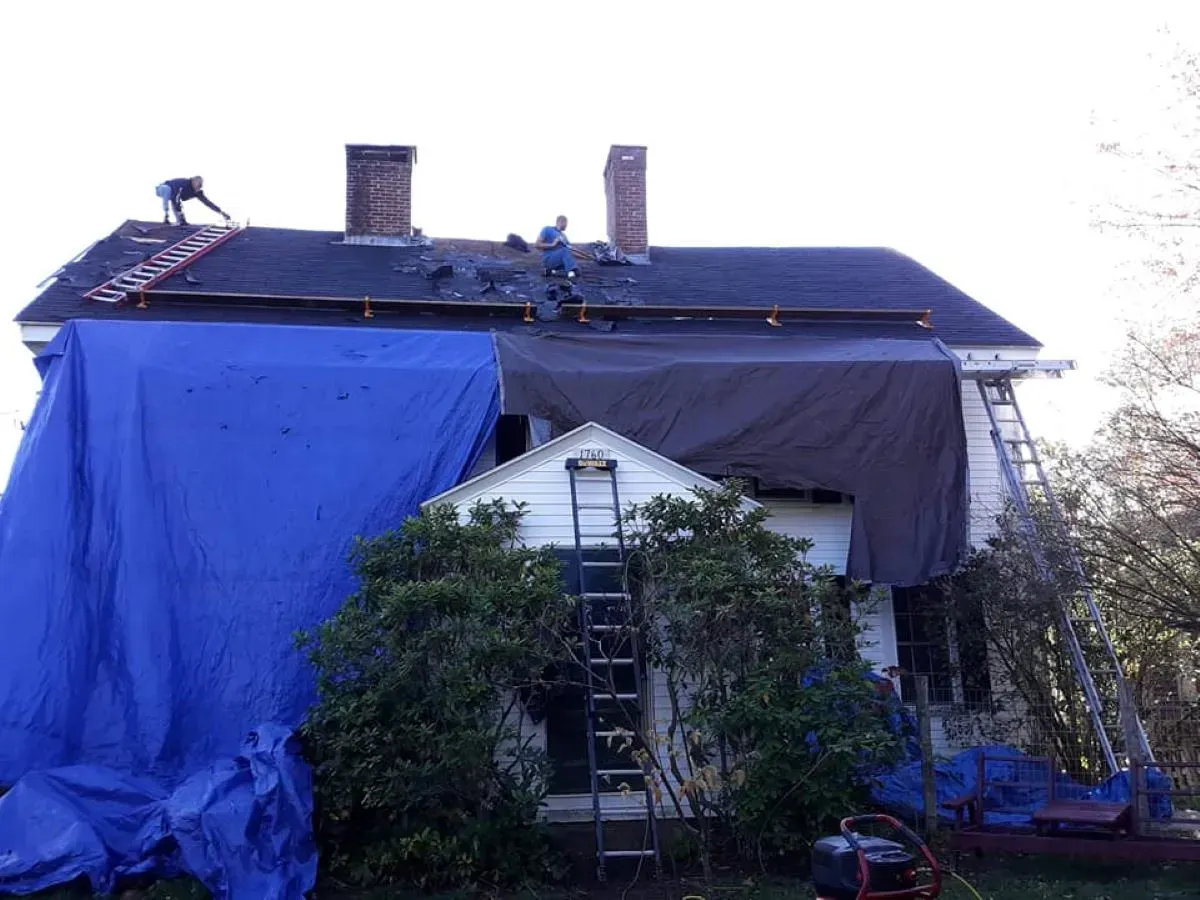 Workers replace a house roof covered by a large blue tarp, using ladders and equipment.