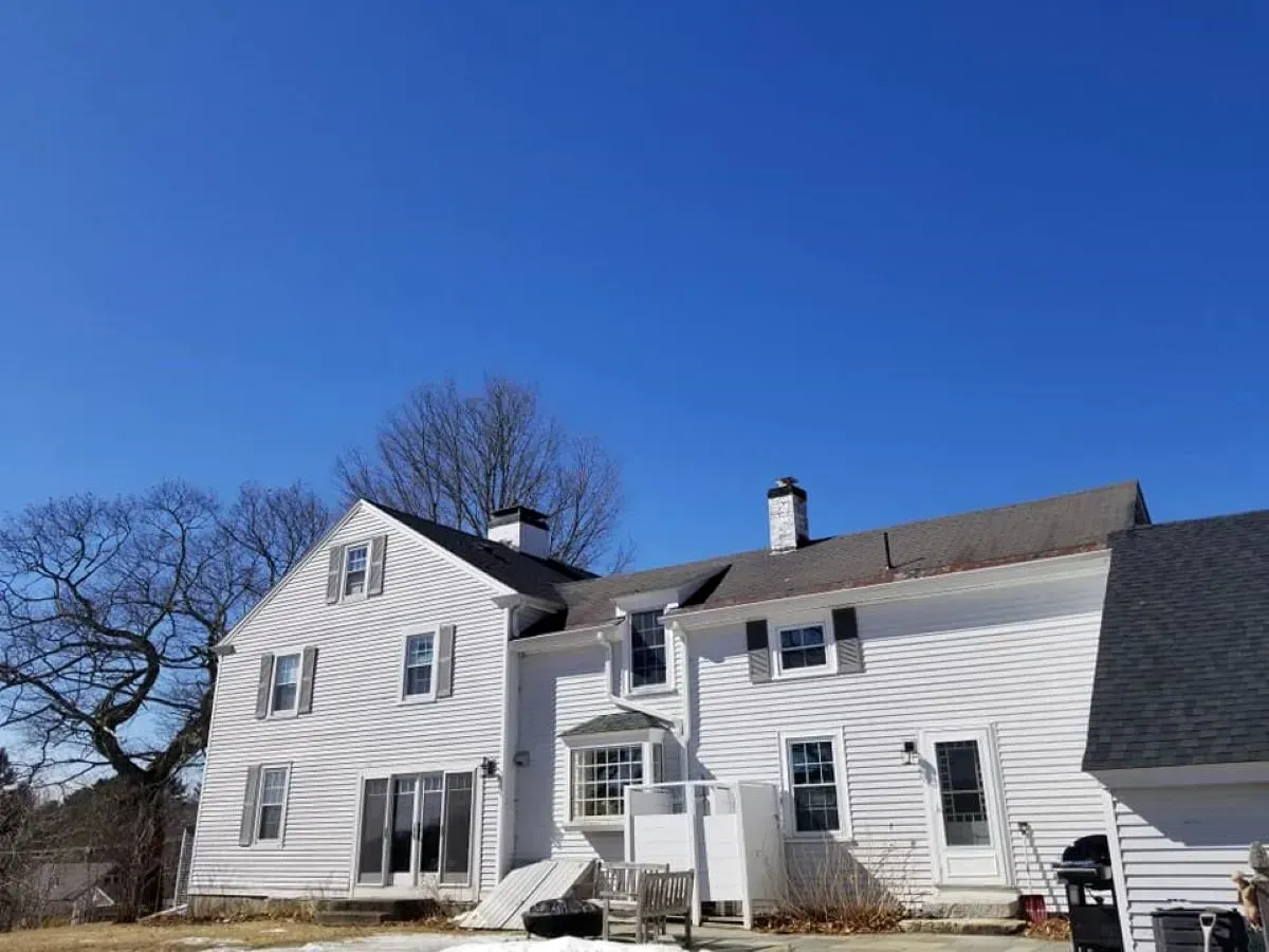 A large, white, two-story house with gray roofing stands against a clear blue sky.