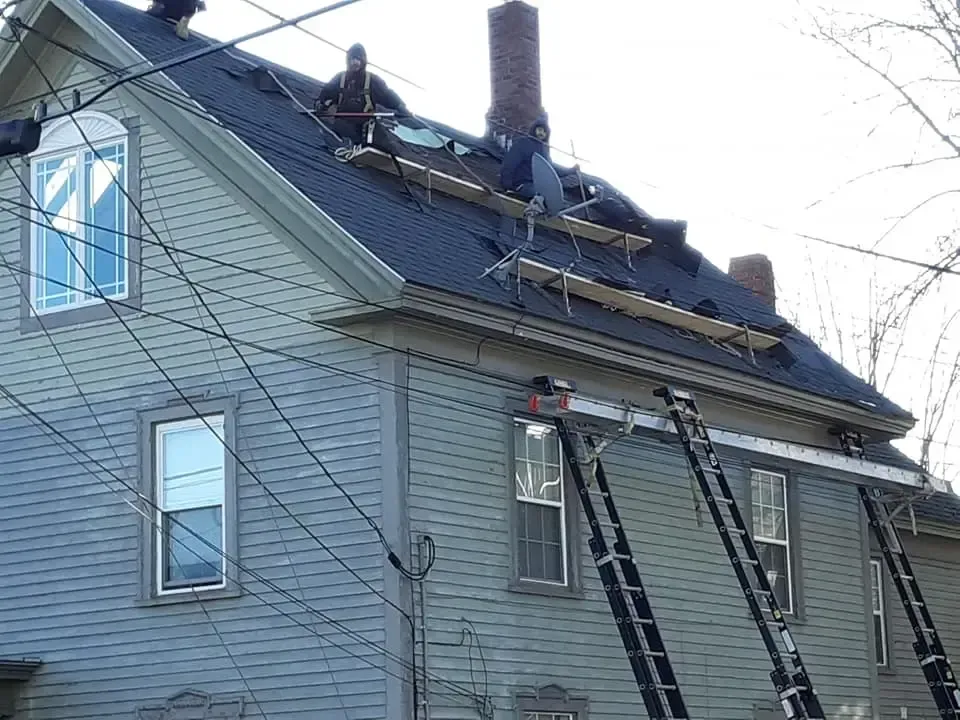 Two roofers working on a partially shingled dark roof of a light blue house with multiple extension ladders.