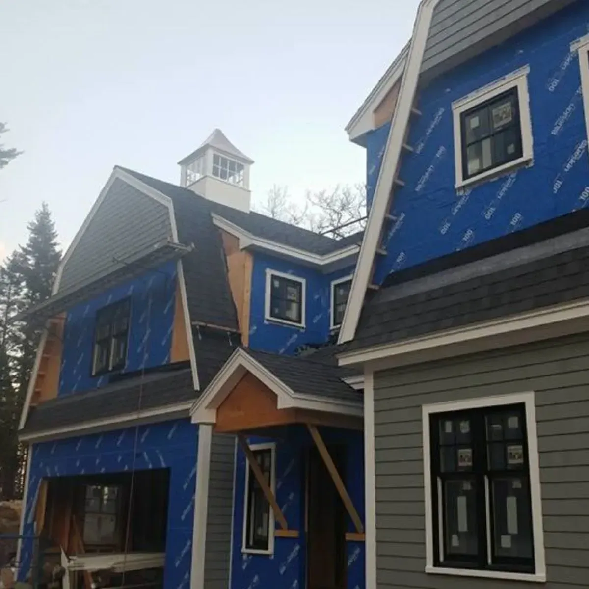 A two-story house under construction shows blue protective wrap and exposed framing, with a small cupola on the roof.