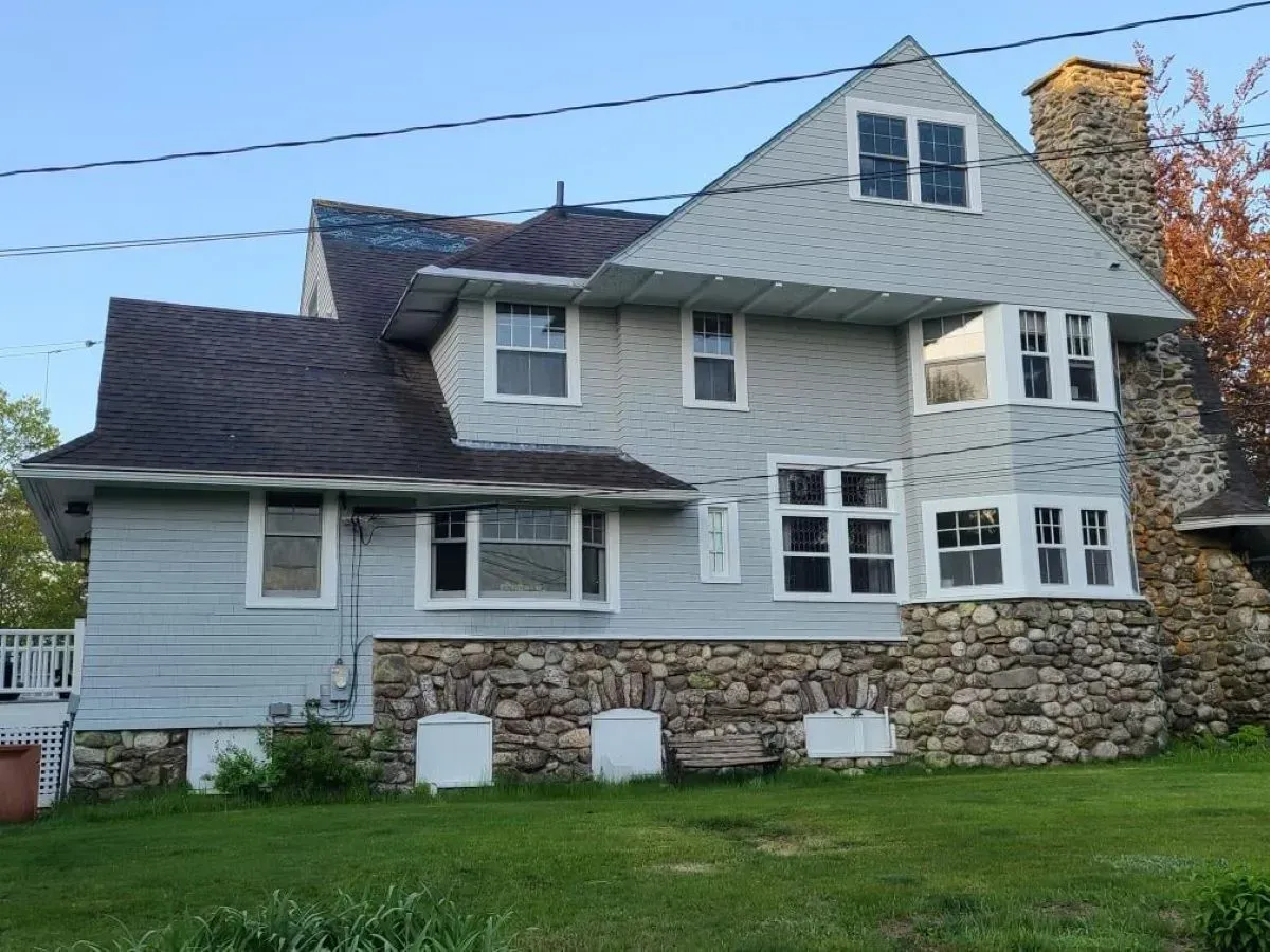 A gray shingle house with a stone base and several windows stands on a grassy lawn.