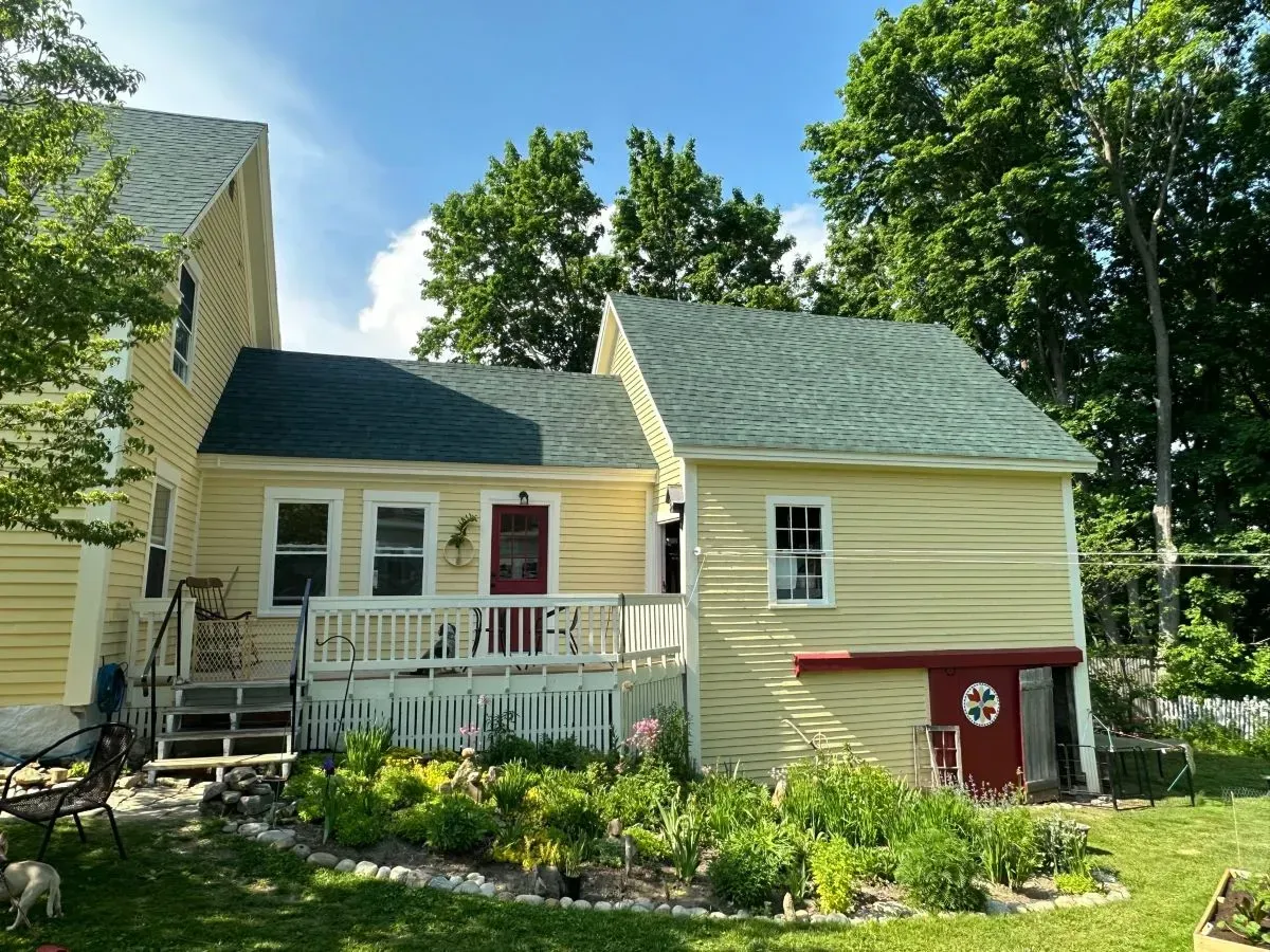 A yellow house with a green roof and white trim sits nestled among lush green trees and a garden.