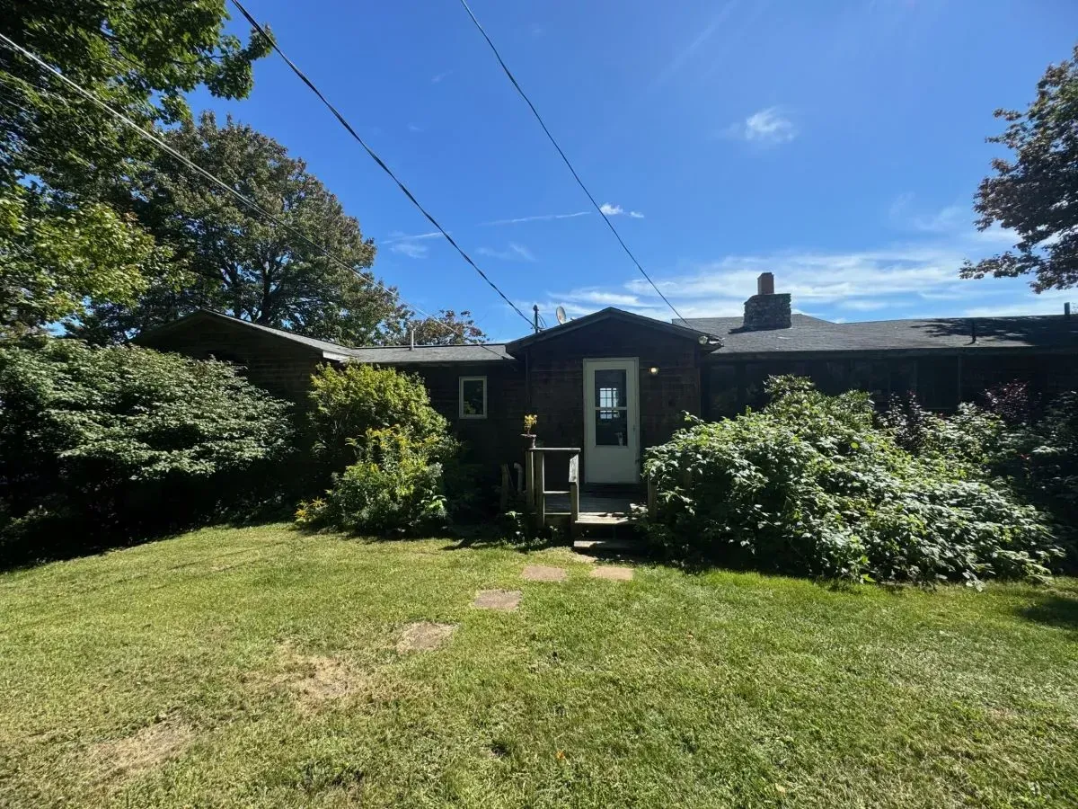 A dark, small house with a single door sits on a grassy lawn under a clear blue sky.