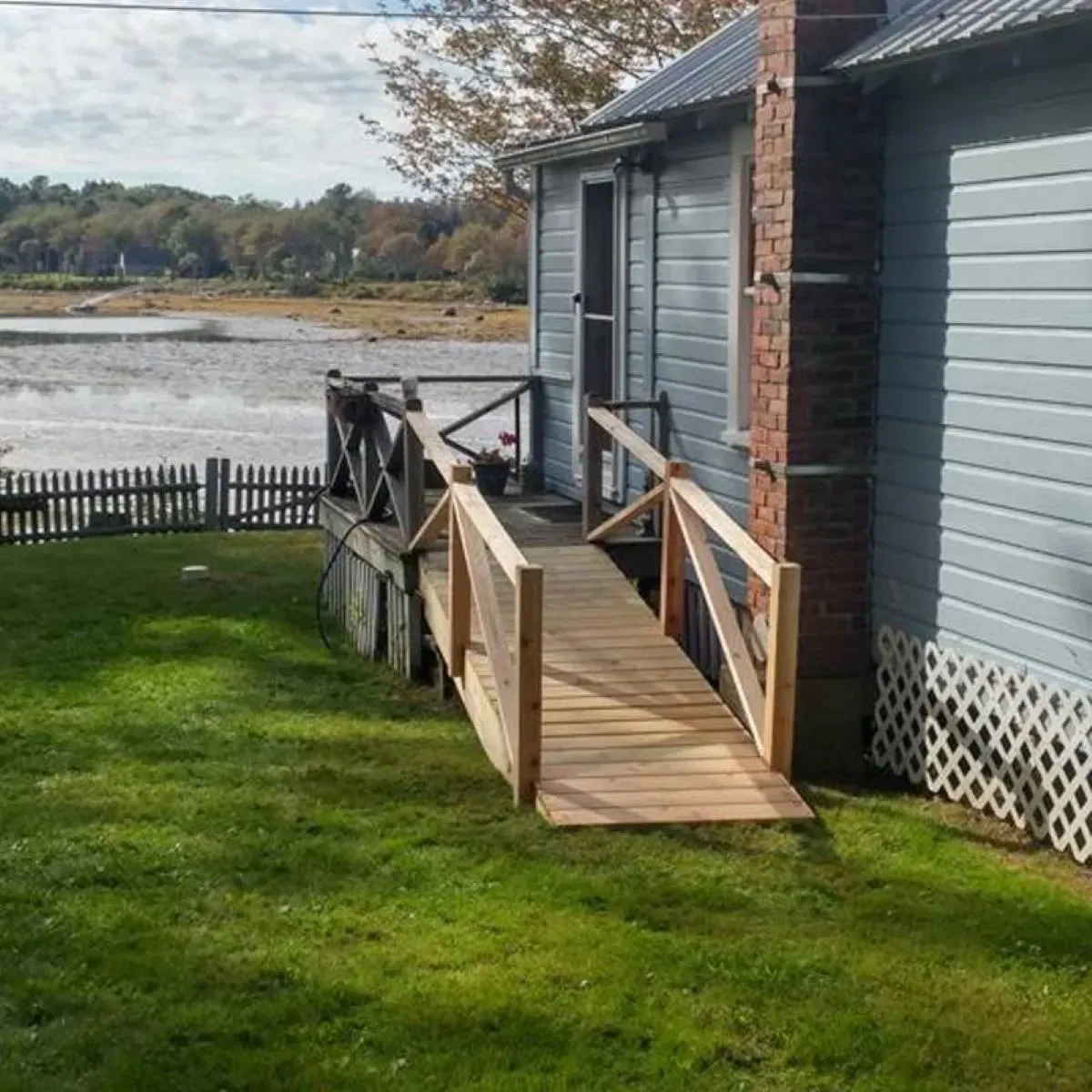 A newly built wooden ramp with handrails leading from a grassy lawn up to a light blue house by grey water.