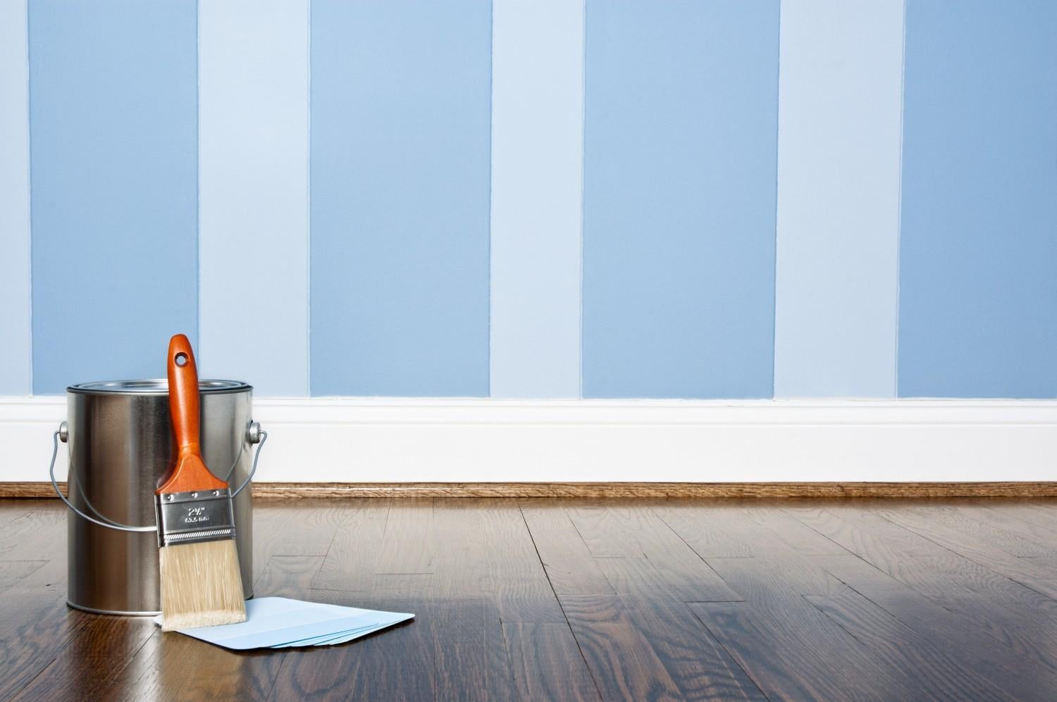 Paint can and brush on a dark wood floor in front of a blue and white striped wall.